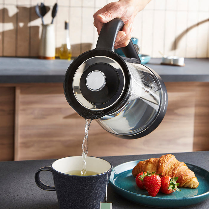 Person pouring tea from a kettle into a mug on a kitchen counter with a plate of croissants and strawberries.