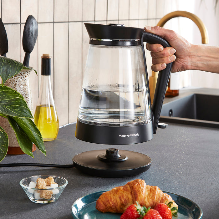 Person holding a black and clear glass electric kettle on a kitchen counter with food and kitchen items in the background.