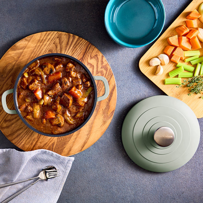 Stew in a pot on a wooden board with vegetables and a lid on a gray surface