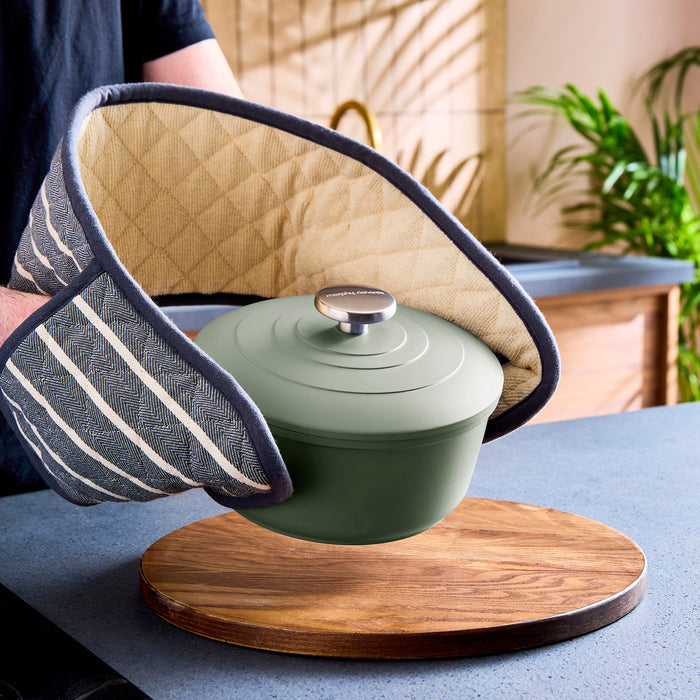 Person holding a green casserole pot with a wooden trivet on a table