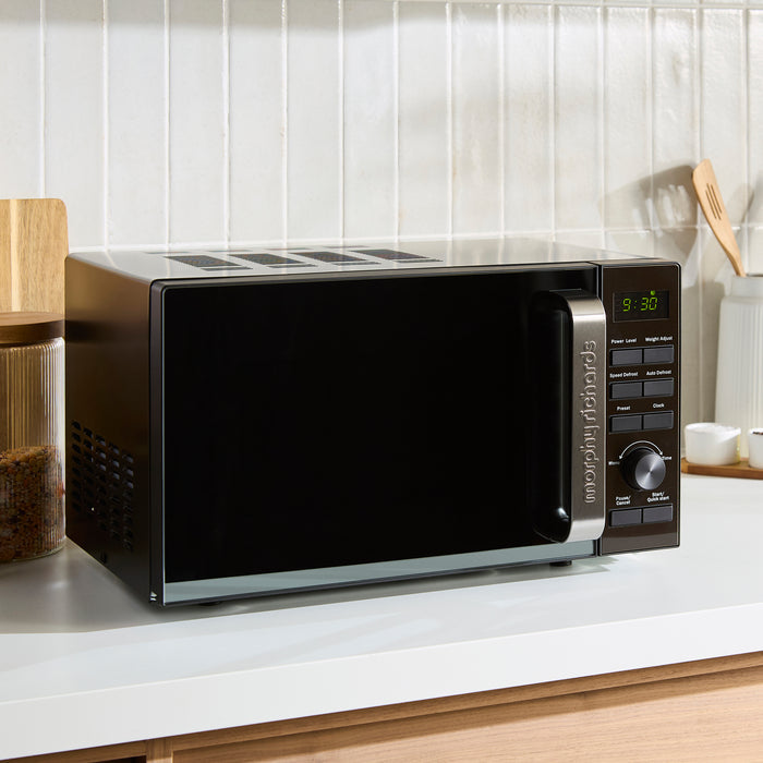 Black microwave oven on a kitchen counter with a white tiled wall in the background.