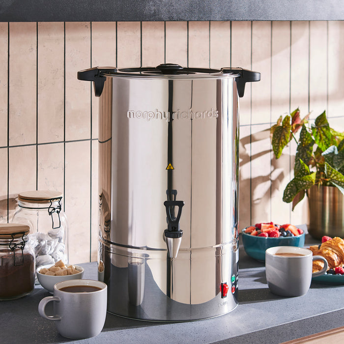 Stainless steel tea urn on a kitchen counter with coffee cups and snacks.