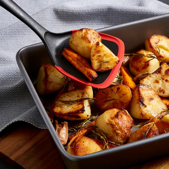 Roasted potatoes with rosemary being scooped out of a baking tray using a red and black spatula.