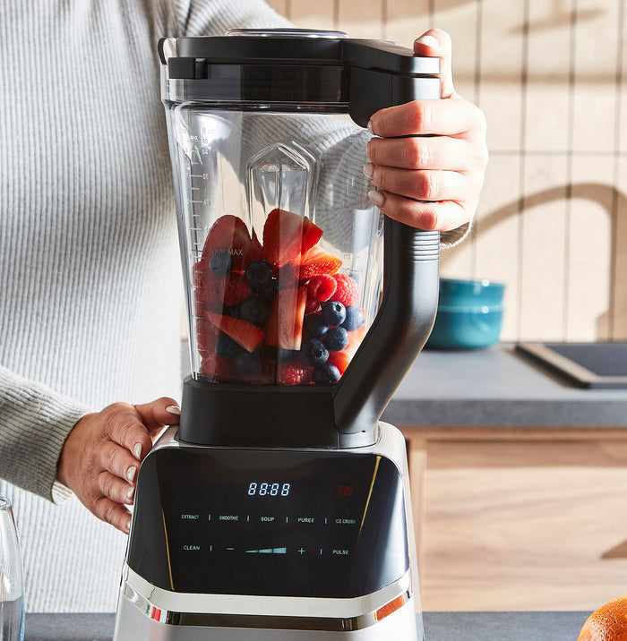 Person holding a blender filled with fruits in a kitchen setting