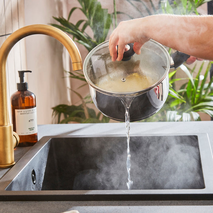 Person pouring water from a pot into a sink with a gold faucet and plants in the background