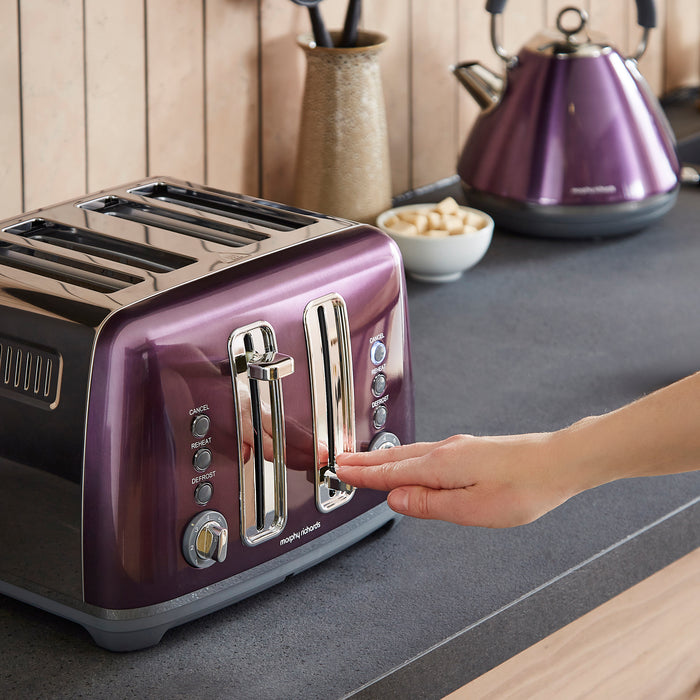 Purple toaster on a kitchen counter with a hand pressing a button, purple kettle in the background.