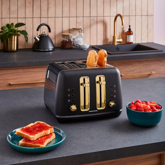 Black toaster with gold accents on a kitchen counter, surrounded by toast and fruit.