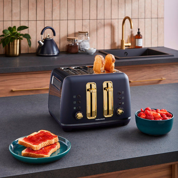 Navy blue toaster with gold accents on a kitchen counter, surrounded by toast, a bowl of fruit, and a kettle.