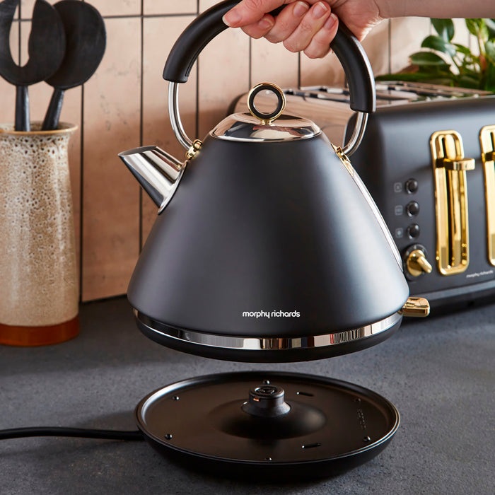 Black Morphy Richards kettle being held above a kitchen counter with a stove and utensils in the background.