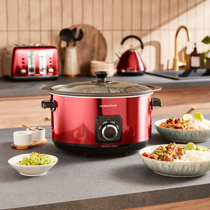 Red slow cooker on a kitchen counter with food and kitchen appliances in the background
