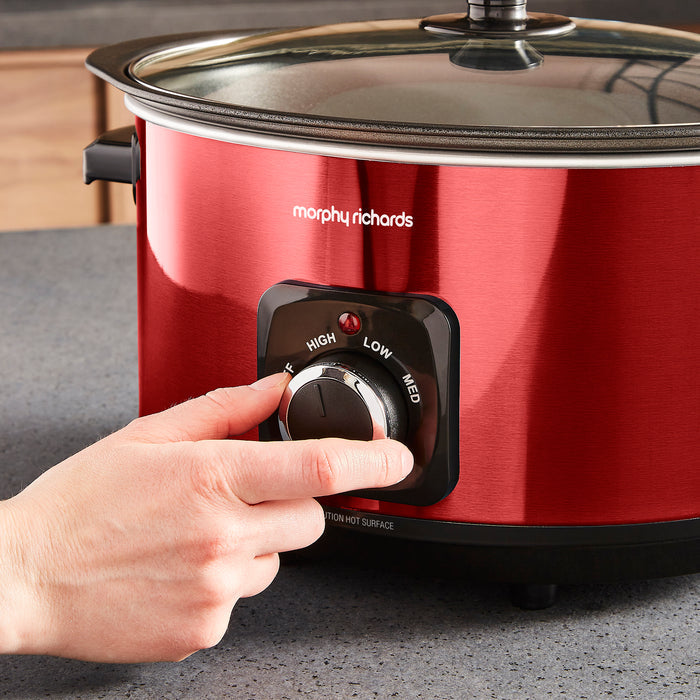 Red Morphy Richards slow cooker with a hand adjusting the dial on a grey surface.