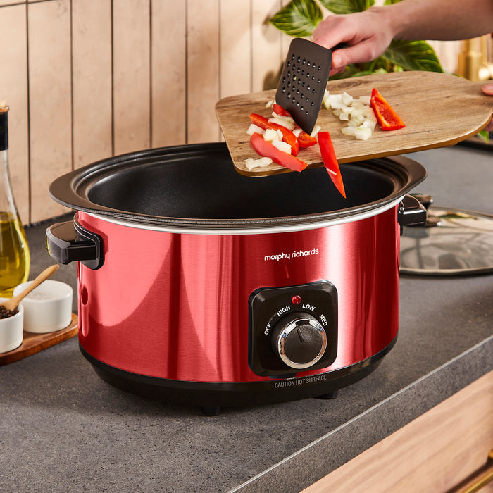 Red Morphy Richards slow cooker with food being added to it in a kitchen setting.
