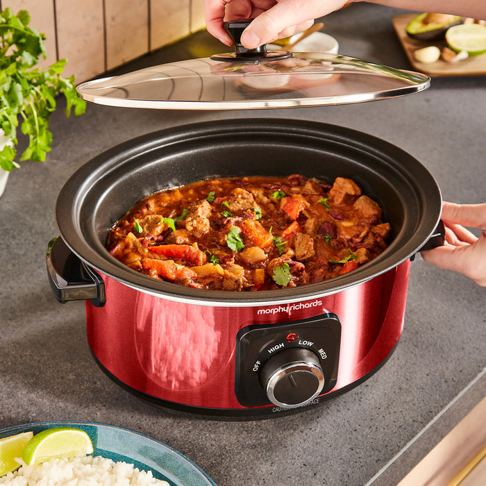 Red Morphy Richards slow cooker with a lid being lifted, revealing food inside on a kitchen counter.