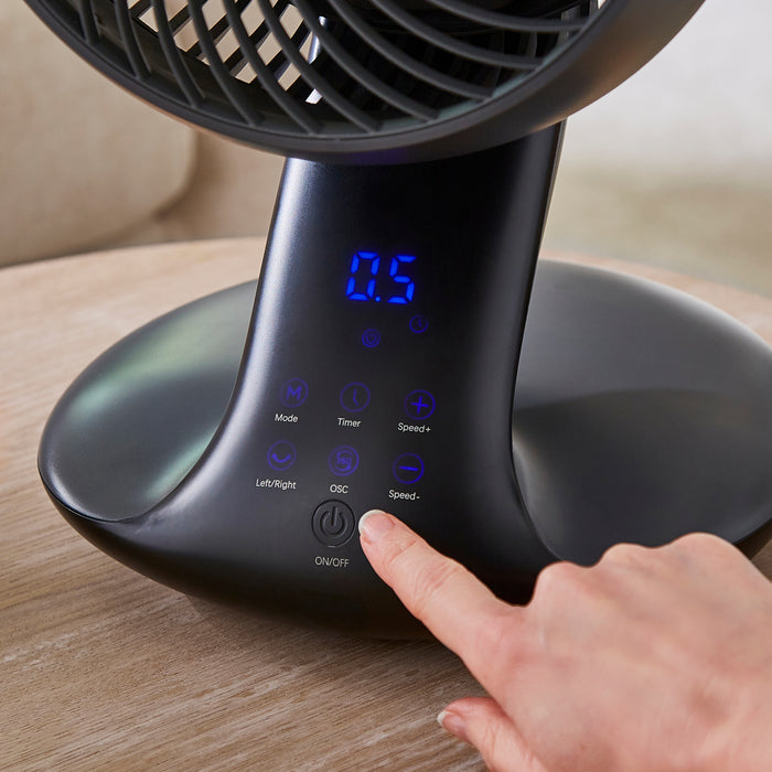 Black fan with digital display and control buttons on a wooden surface, with a hand pressing a button.