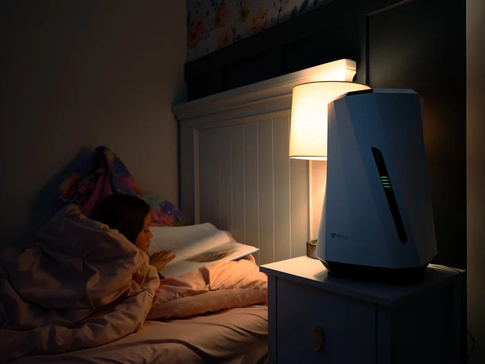 Person lying in bed with an Airdog humidifier on a nightstand next to a lamp in a dark room