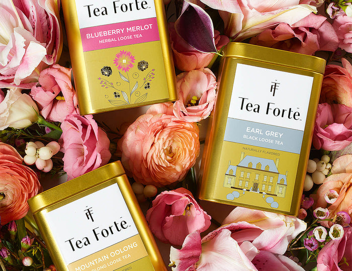 Three gold tea tins from Tea Forté surrounded by pink and orange flowers.
