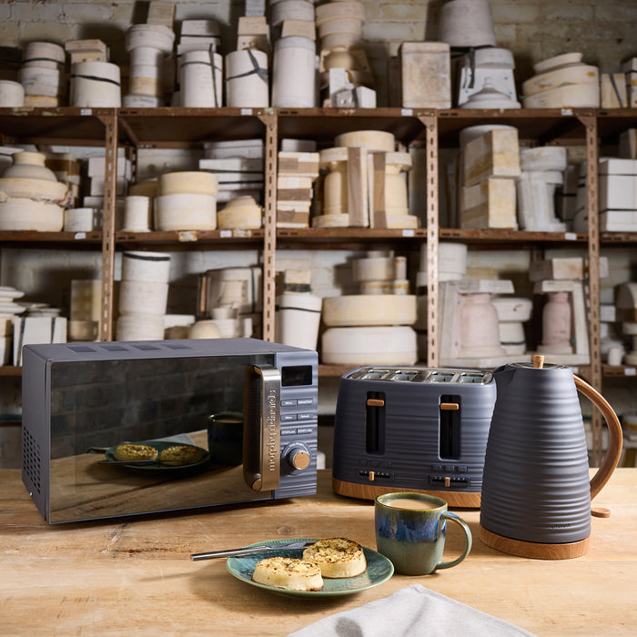 Kitchen appliances including a microwave, toaster, and kettle on a wooden table with shelves of kitchenware in the background.