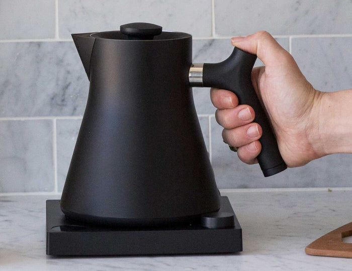 Hand picking up the Black Corvo Kettle EKG Pro by its handle as it rests on it's base in a kitchen.