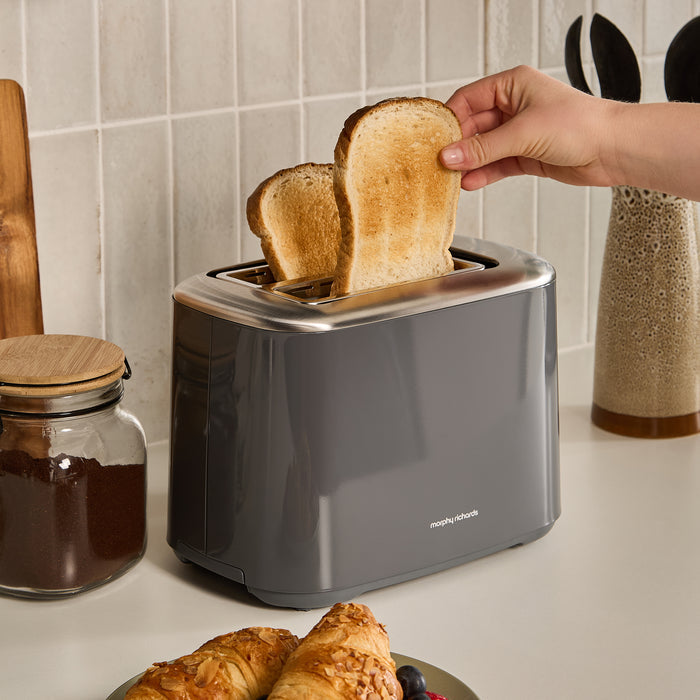 Person taking toast out of a 2 slice toaster with a kitchen setting in the background.