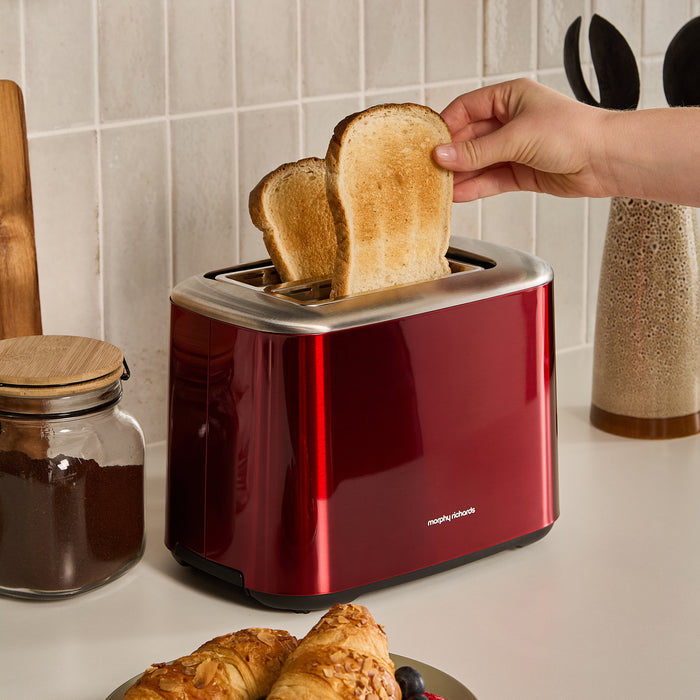 Red toaster with slices of toast being removed, next to a jar of jam and a plate of pastries.