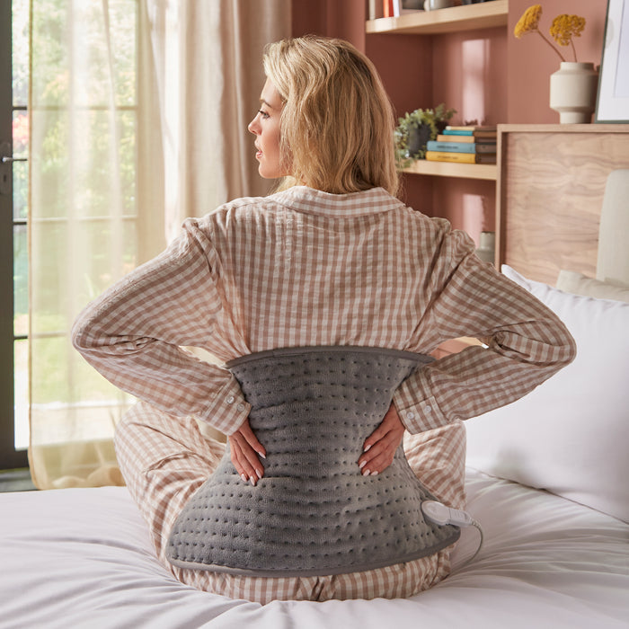 Woman sitting on a bed using a heating pad in a cosy living room.