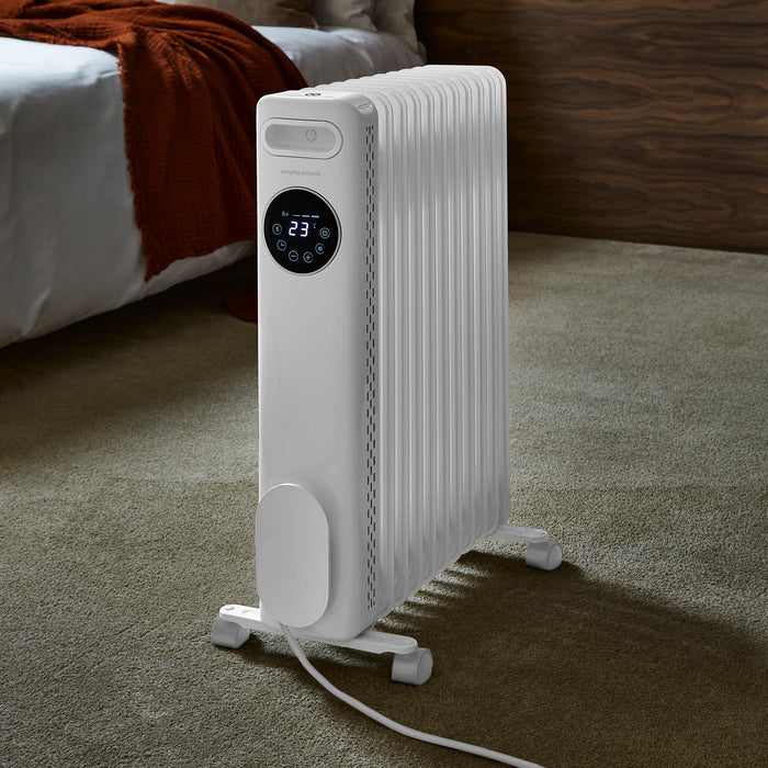 White electric radiator with digital display on a carpeted floor, bedroom setting.