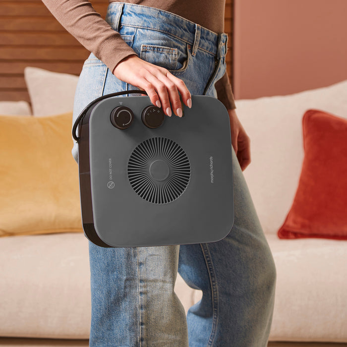 Person holding a portable grey heater in a living room setting