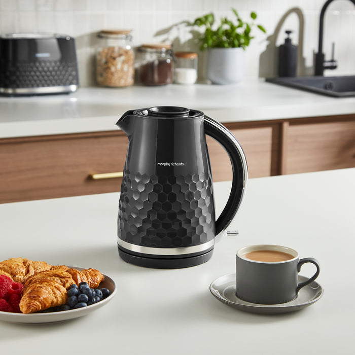 Black electric kettle on a kitchen counter with a plate of food and a cup of coffee.