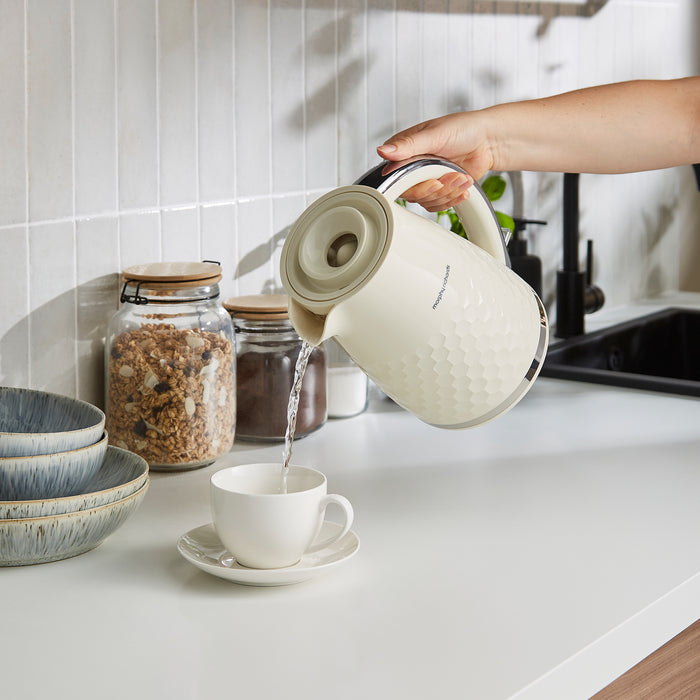 Person pouring water from a white ceramic kettle into a white mug on a kitchen counter.