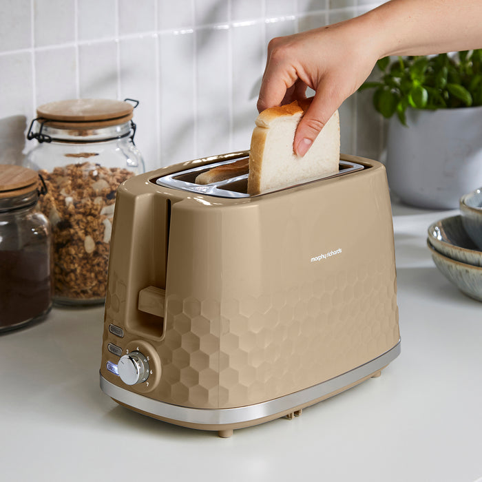 A beige toaster with a honeycomb texture sits on a white countertop in a kitchen setting, with a hand inserting a slice of white bread into the top slot.