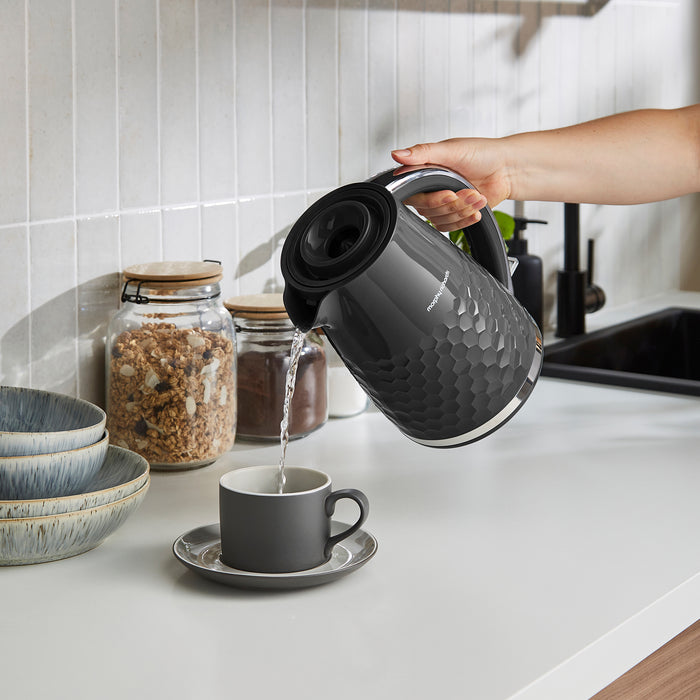 Person pouring water from a black electric kettle into a gray mug on a kitchen counter.