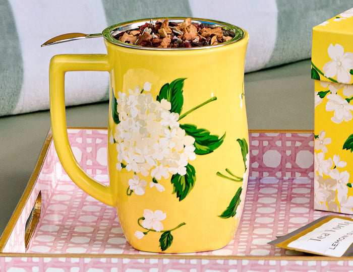 Yellow steeping cup with white hydrangea flowers and green leaves, filled with loose tea and metal infuser spoon.
