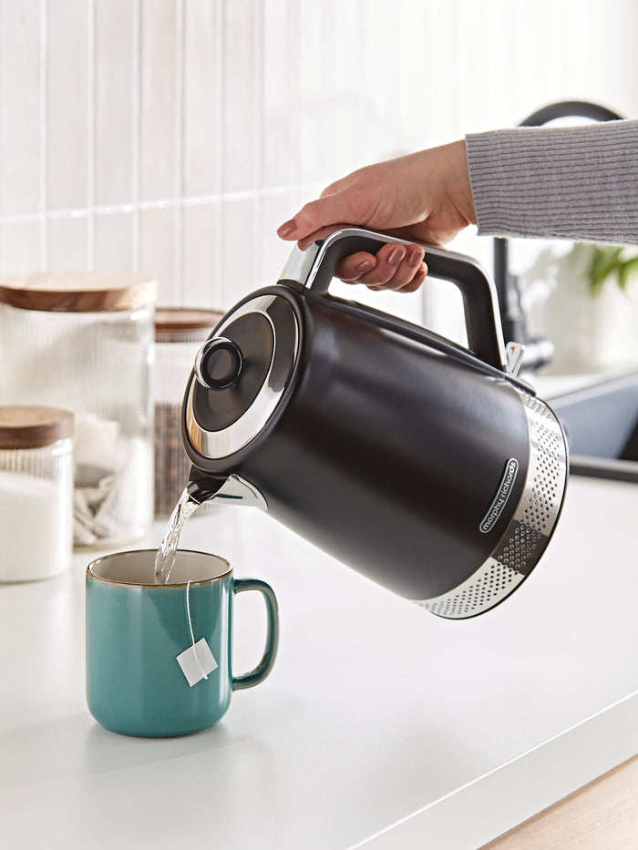 Person pouring water from a black electric kettle into a teal mug on a kitchen counter.