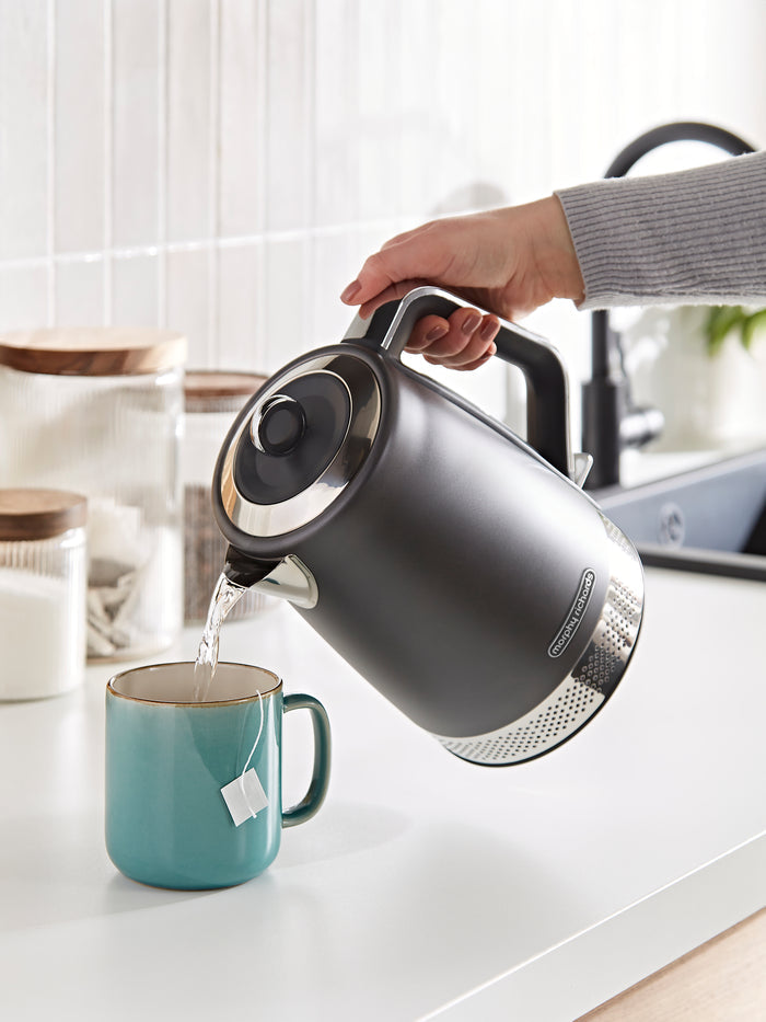 Person pouring water from a gray electric kettle into a blue mug on a kitchen counter.