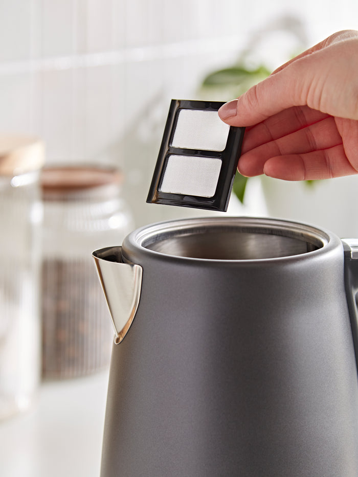 Hand placing a filter into a silver kettle on a blurred kitchen background