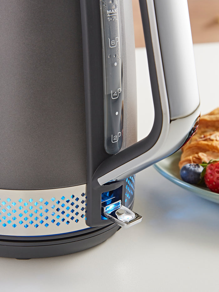 Gray electric kettle with blue accents on a white surface, next to a plate of food.