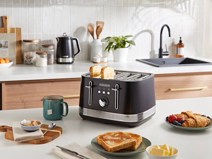 Black toaster on a kitchen counter with toast, mugs, and a plate of food.