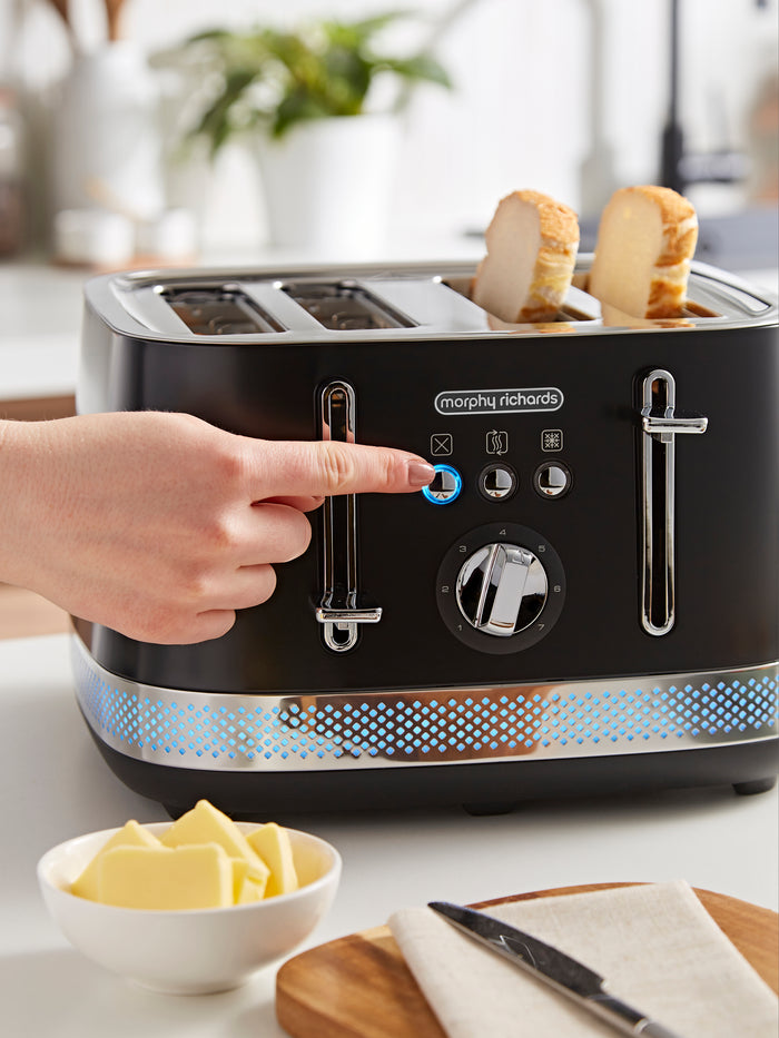 Black Morphy Richards toaster with four slices of bread, a hand pressing a button, and a bowl of butter on a kitchen counter.