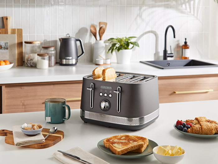 Gray toaster on a kitchen counter with toast, mugs, and fruit plate.