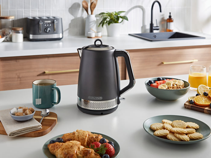 Black electric kettle on a kitchen counter with breakfast items and appliances in the background.