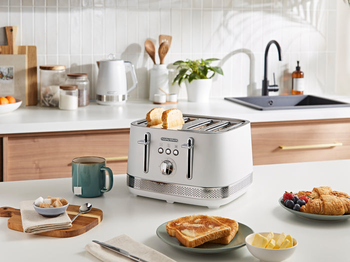 White toaster on a kitchen counter with bread slices, a mug, and a plate of food.