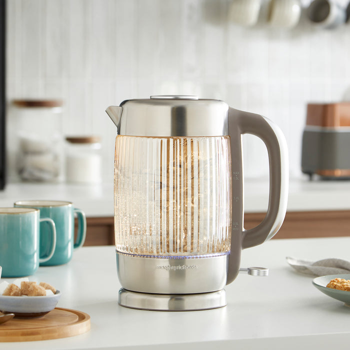 Clear glass electric kettle on a kitchen counter with mugs and kitchen items in the background