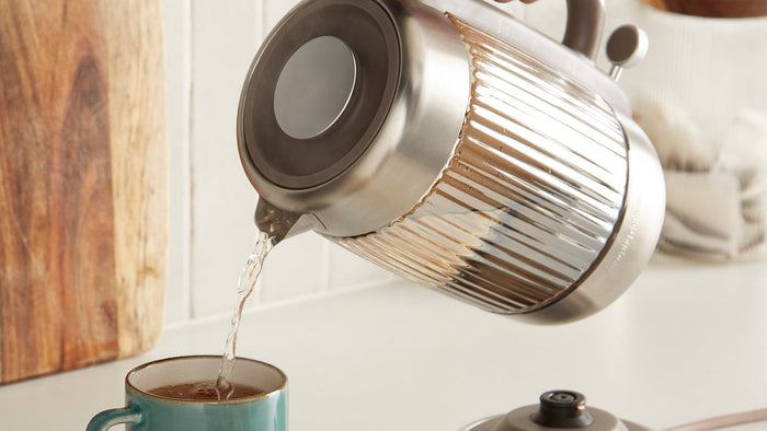 Silver kettle pouring liquid into a teal mug on a white surface with a wooden wall in the background.