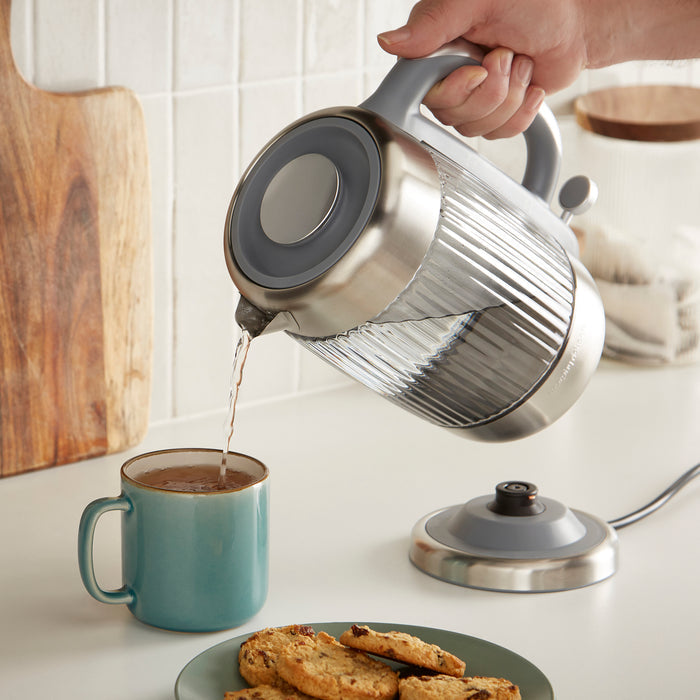 Person pouring water from a silver electric kettle into a blue mug on a kitchen counter.