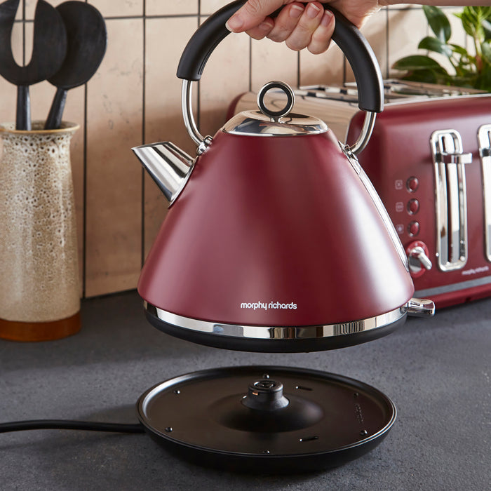 Red kettle with black handle held by a hand on a kitchen counter, with a Morphy Richards logo.