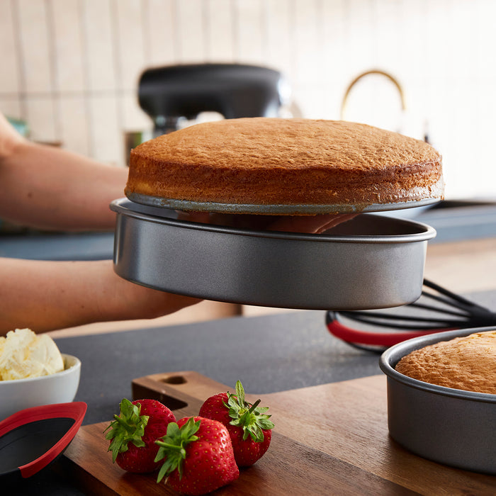 Baked cake in a round metal baking pan on a kitchen counter with strawberries and cream.