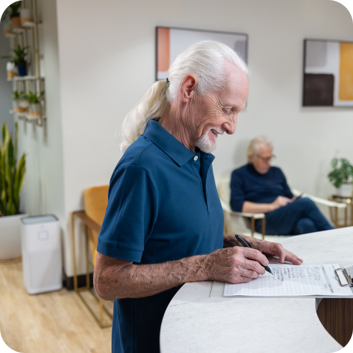 A patient smiling filling out paper work at a doctors office  filled with chairs, plants and a air purifier.