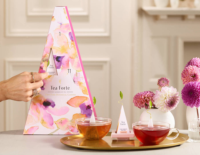 MAIN PDP | Mariposa Advent - 12 Days of Blissful Teas with a hand opening one of the doors on a table with glasses of tea and pink zinnias.