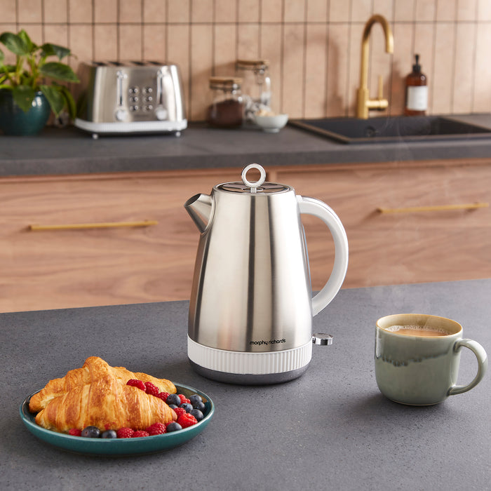Stainless steel kettle on a kitchen counter with a plate of croissants and berries, and a cup of coffee.