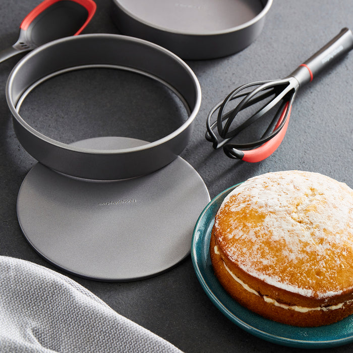 Baking pans, a cake, and a whisk on a dark surface